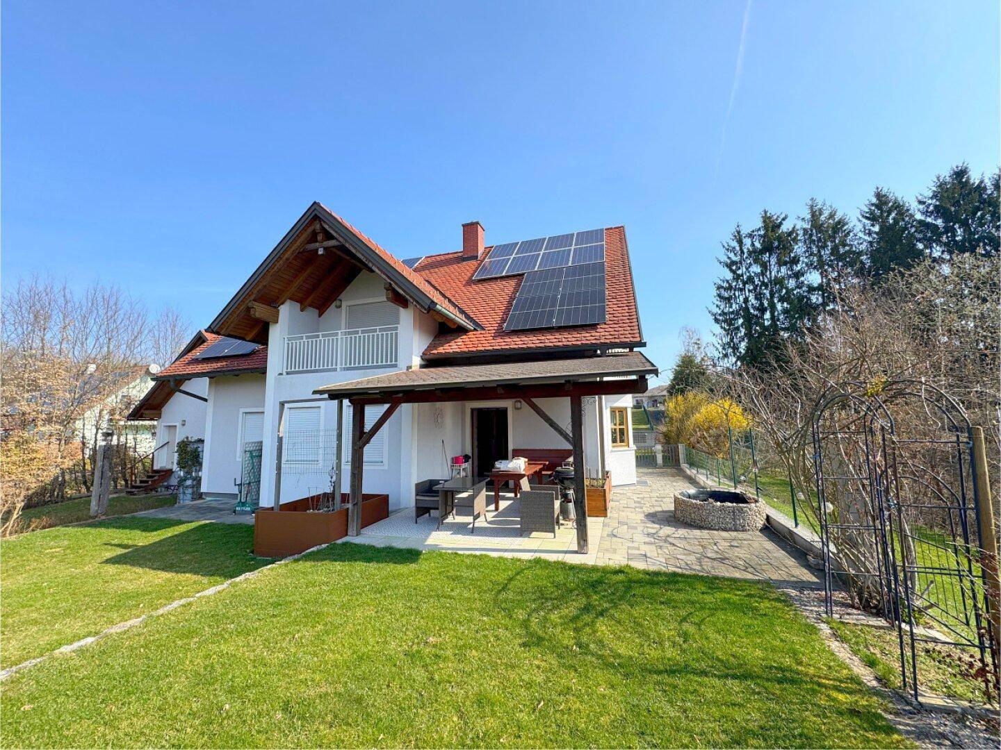 Rückseite des Hauses mit überdachter Terrasse, Gartenmöbeln, Feuerplatz und weitläufigem Rasenbereich.