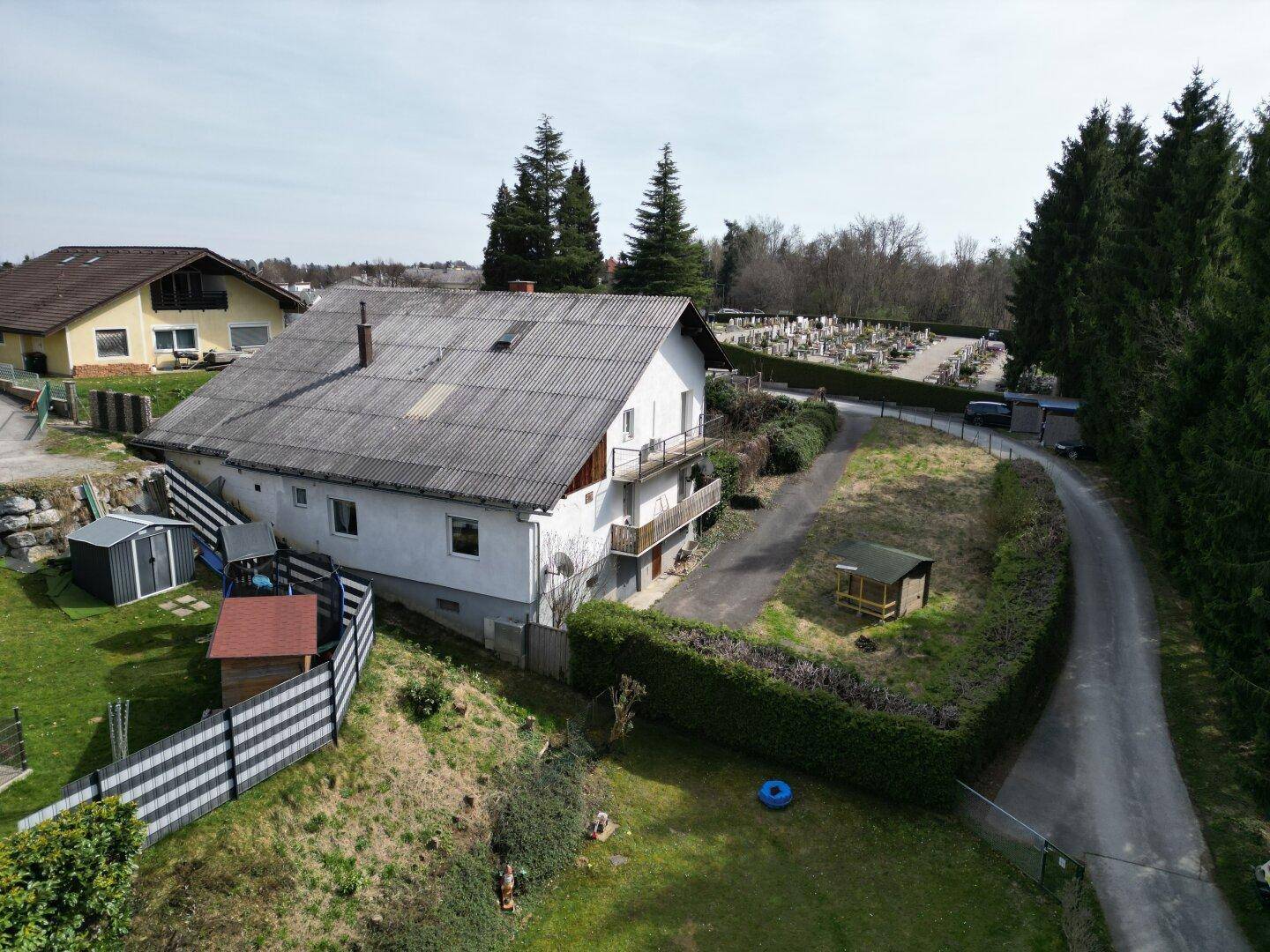 Rückansicht des Hauses mit Gartenbereich, Zufahrt und angrenzendem Friedhof unter blauem Himmel.