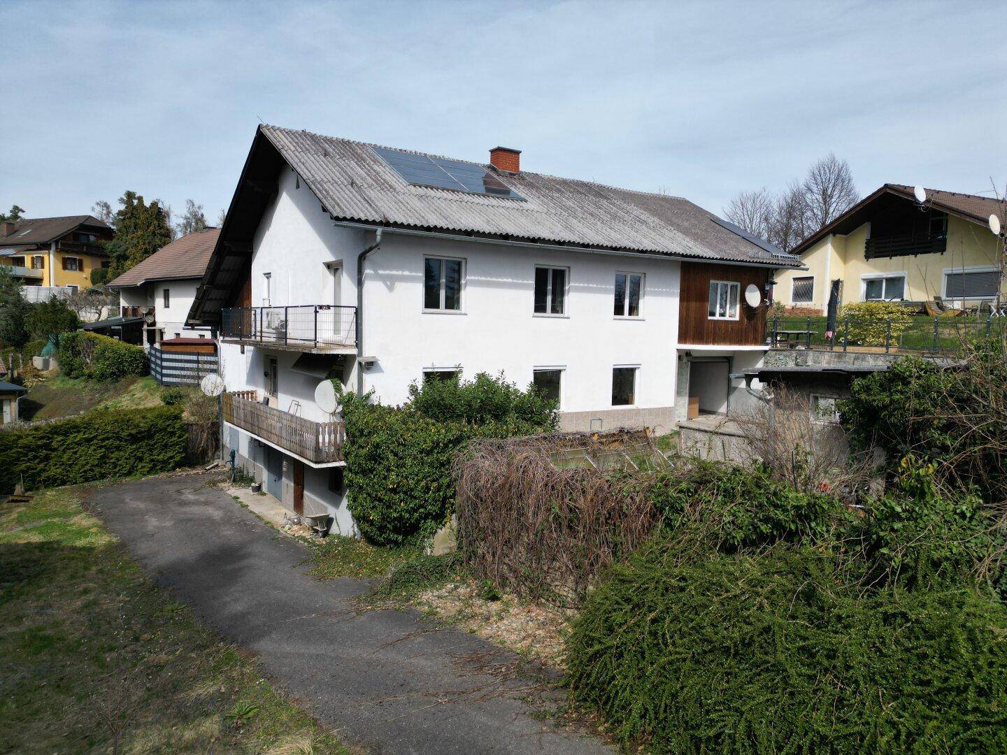 Wohnhaus mit weißer Fassade, zwei Balkonen und Photovoltaikanlage unter blauem Himmel und viel Grün.