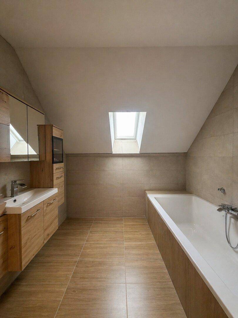 Modernes Badezimmer im Dachgeschoss mit Badewanne, Holzoptik-Möbeln und Dachfenster.