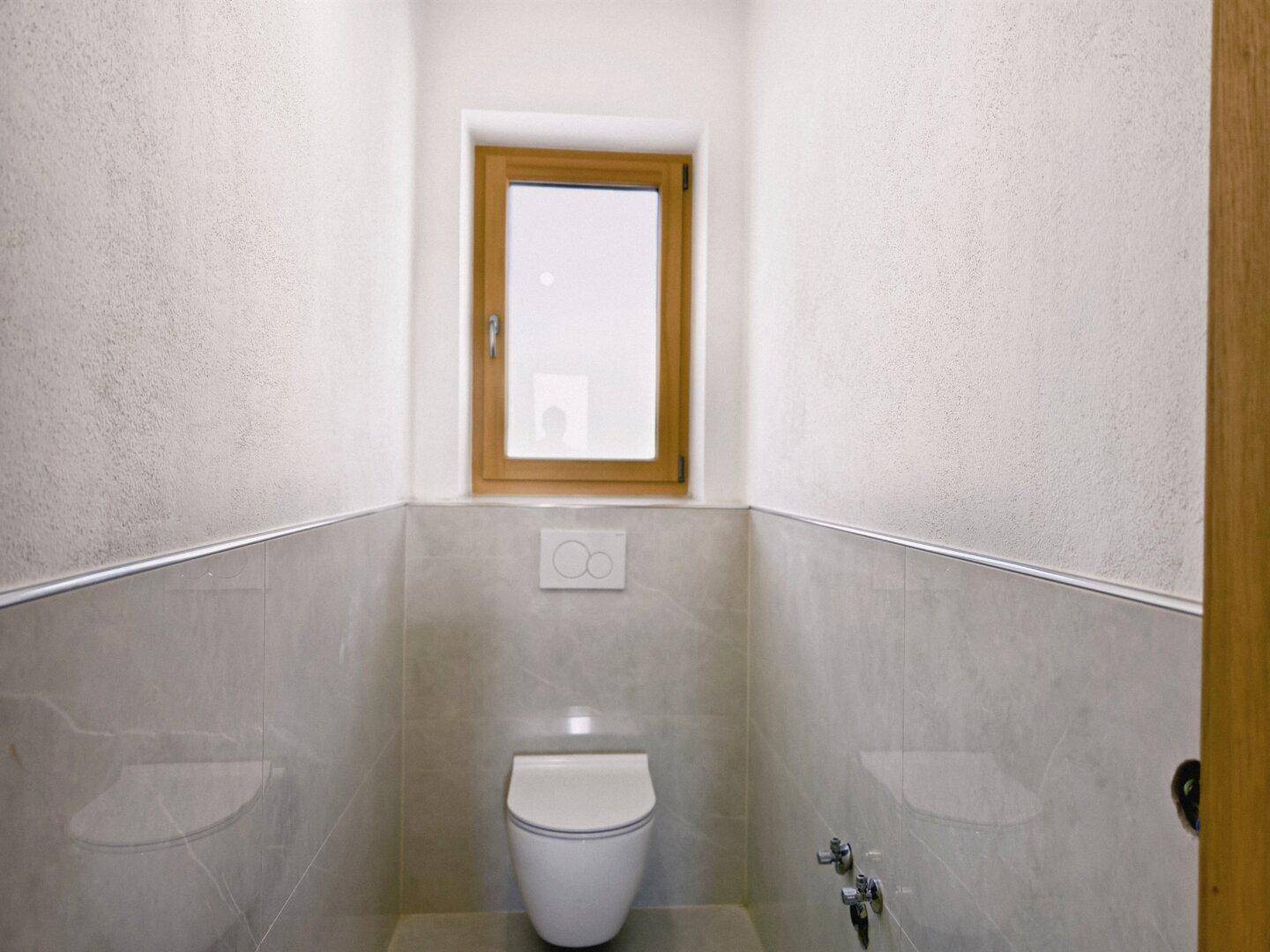 Modernes Gäste-WC mit Wand-WC, Fliesen und kleinem Fenster für natürliche Belüftung.