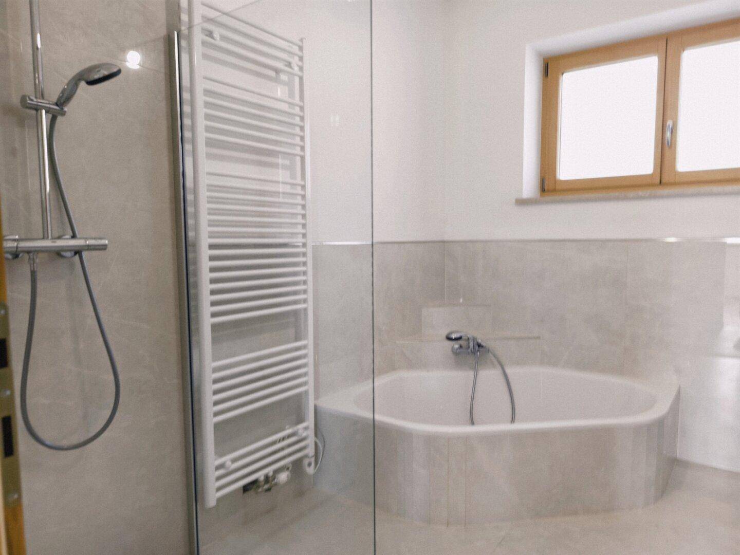 Helles Badezimmer mit Badewanne, Dusche, beheiztem Handtuchhalter und Fenster.