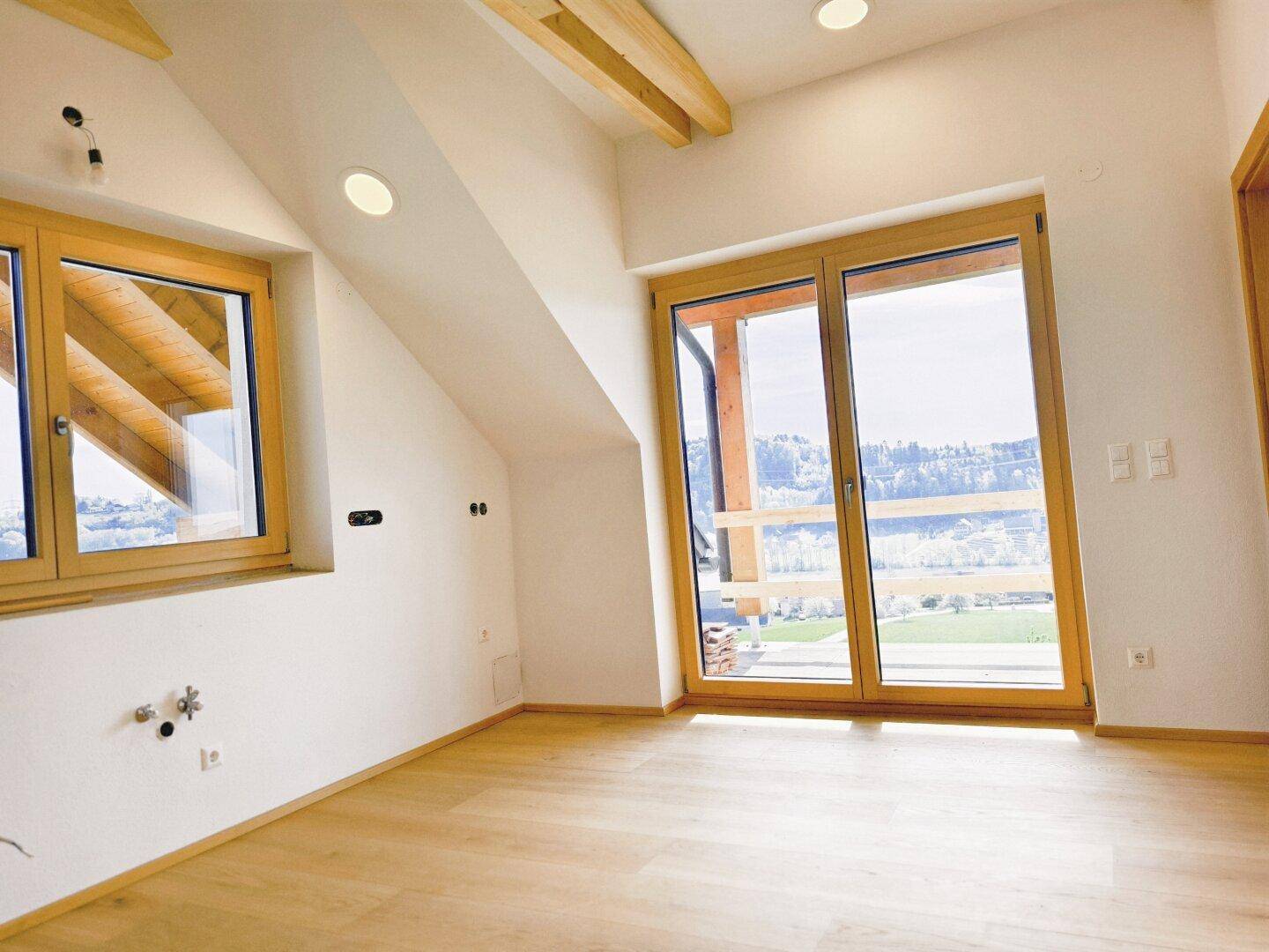 Heller Dachgeschossraum mit Holzboden, sichtbaren Holzbalken und großen Fenstern mit Ausblick.