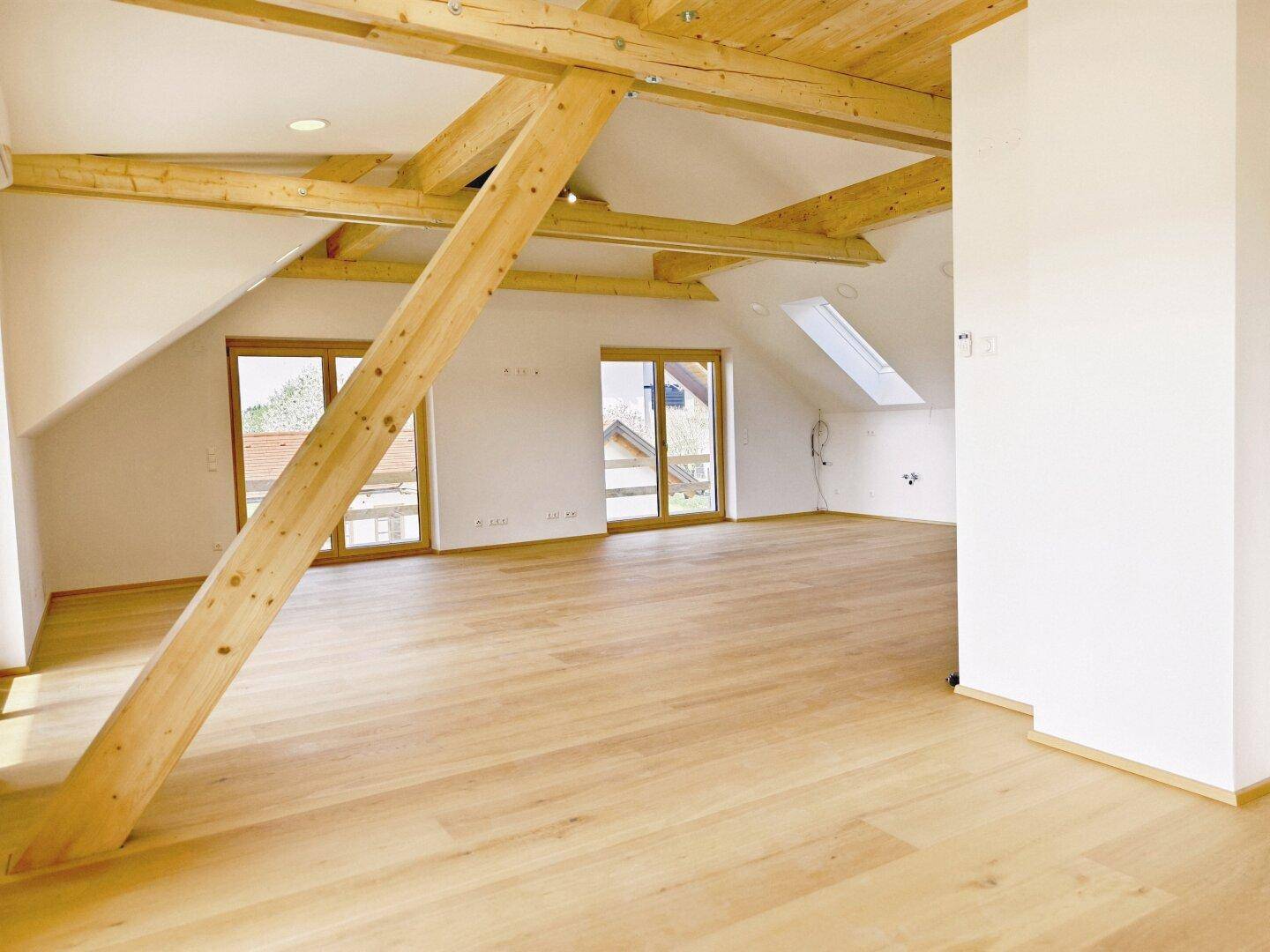 Offener Dachgeschossraum mit Holzboden, sichtbaren Holzbalken und großen Fenstern mit Ausblick.
