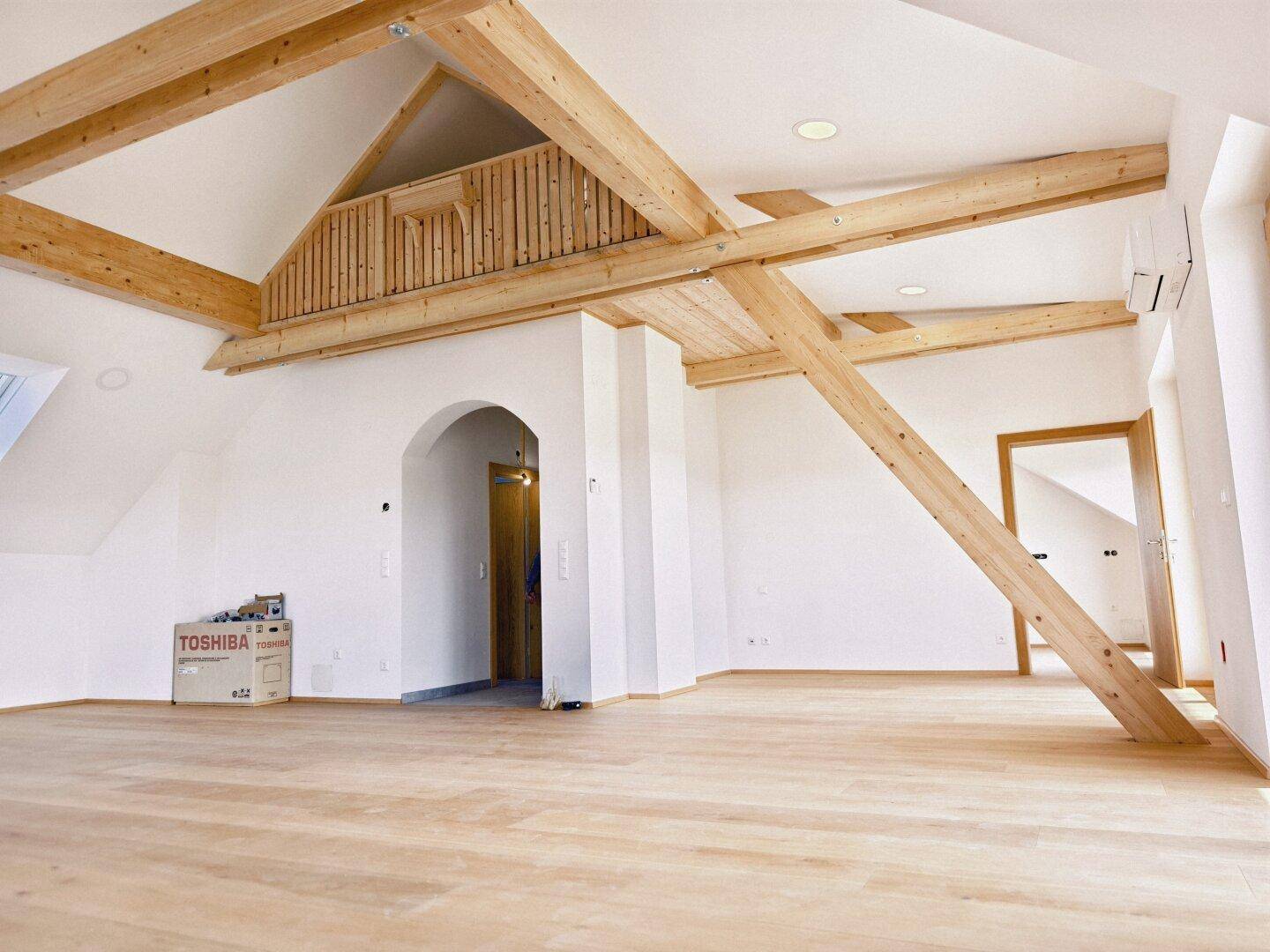 Großzügiger Dachgeschossraum mit sichtbaren Holzbalken, Holzboden und offener Gestaltung.