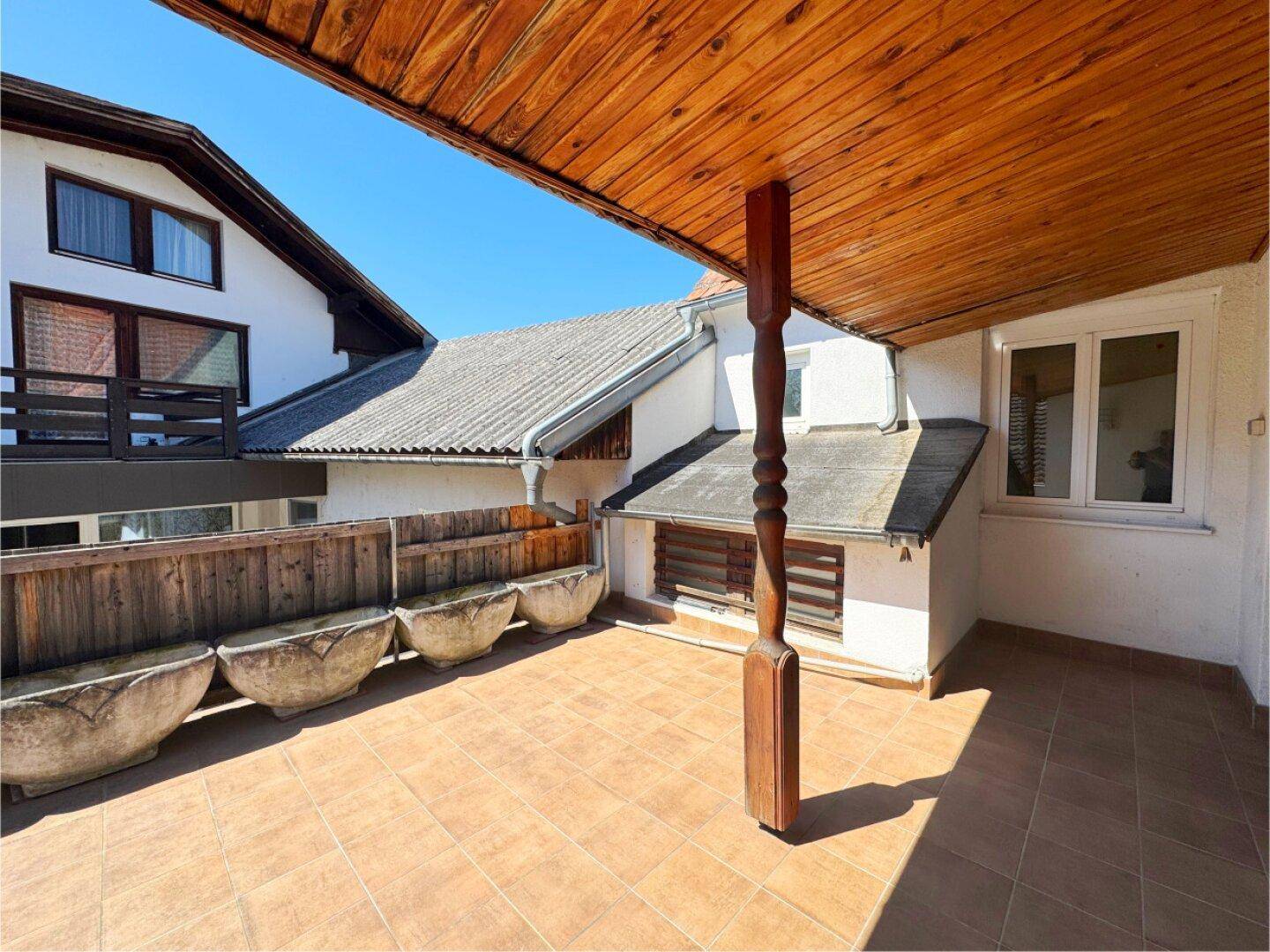 Geräumige Terrasse mit Holzdach, Fliesenboden und Blick auf die umliegenden Dächer und den blauen Himmel.