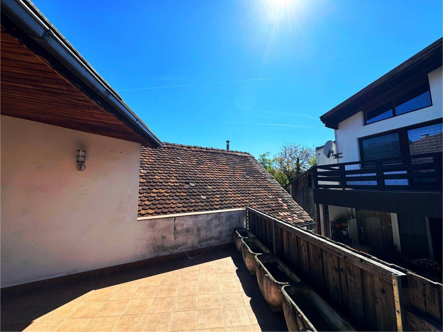 Sonniger Terrassenbereich mit Fliesenboden und Blick auf die Dächer der Nachbargebäude und den klaren Himmel.