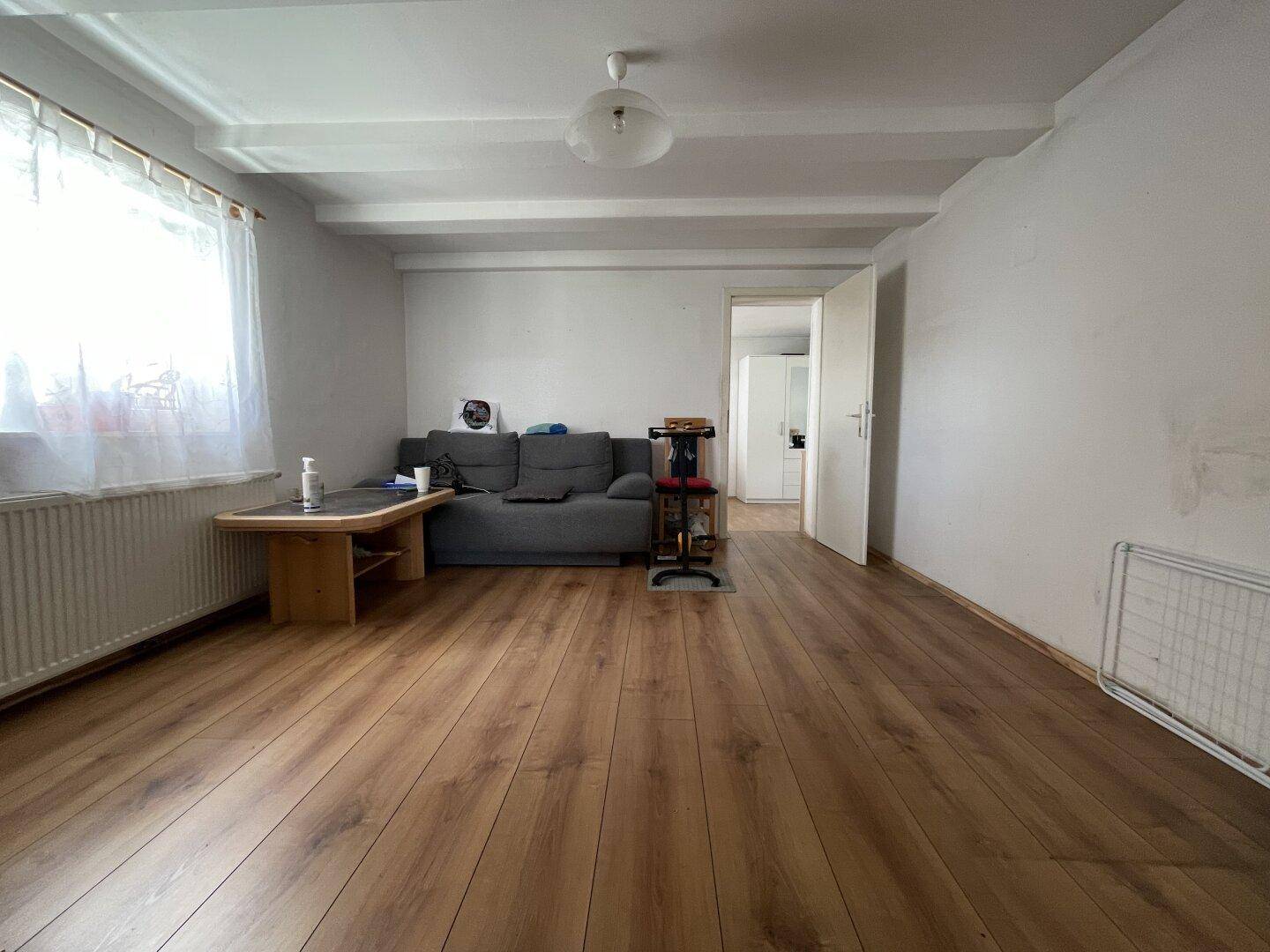 Helles Wohnzimmer mit grauem Sofa, Holzboden und sichtbaren Deckenbalken, bietet viel natürliches Licht.