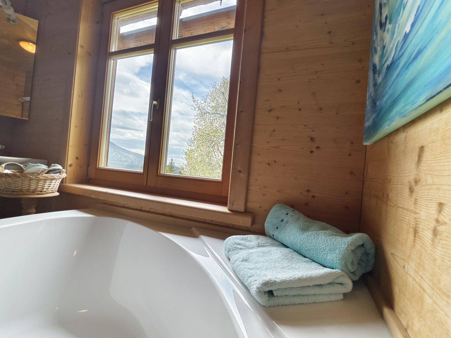 Badezimmer mit Badewanne, Holzvertäfelung und Fenster mit Blick auf die grüne Landschaft.