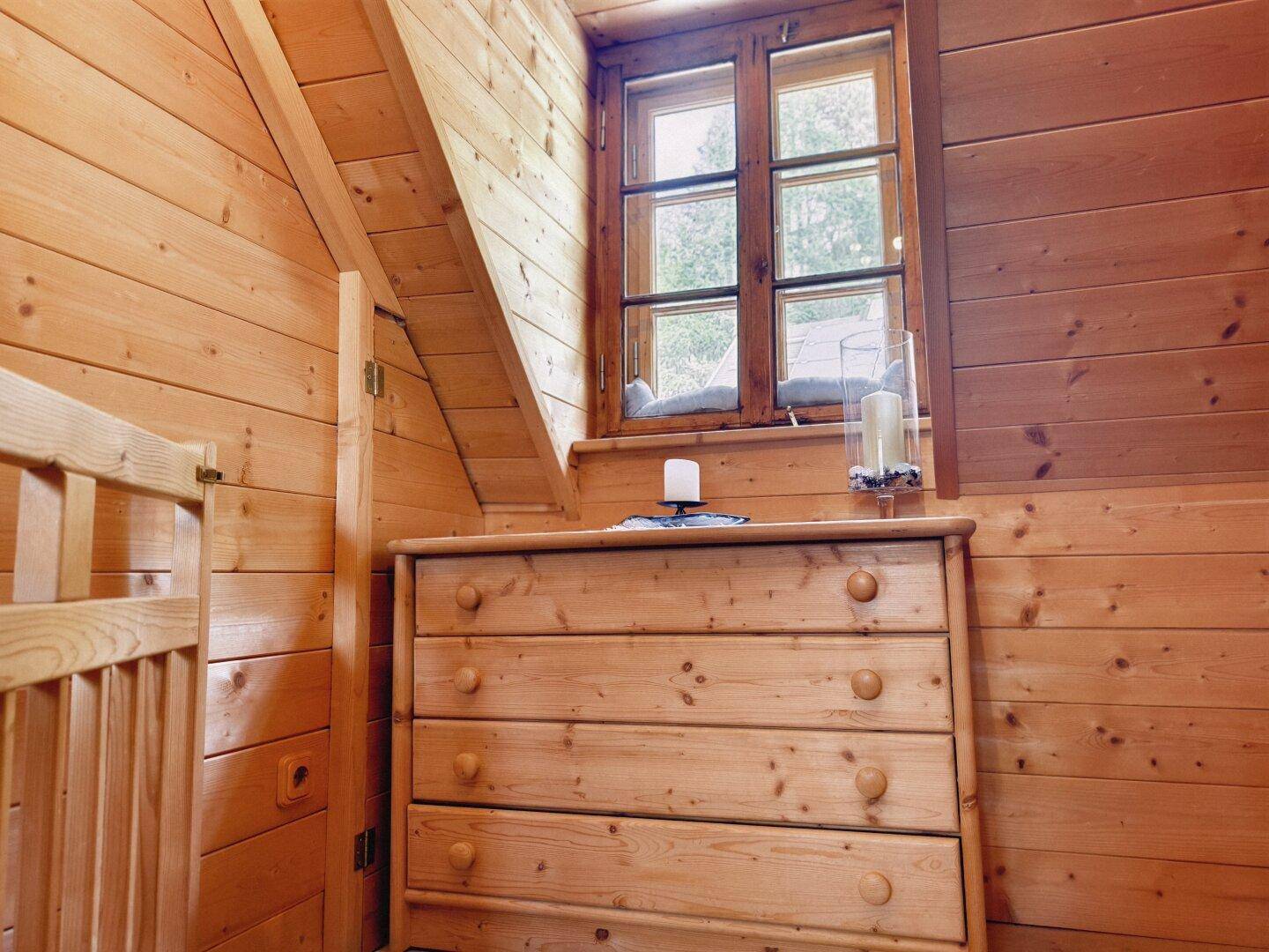 Kleiner, holzvertäfelter Raum im Dachgeschoss mit Kommode und Fenster, das viel Licht hereinlässt.