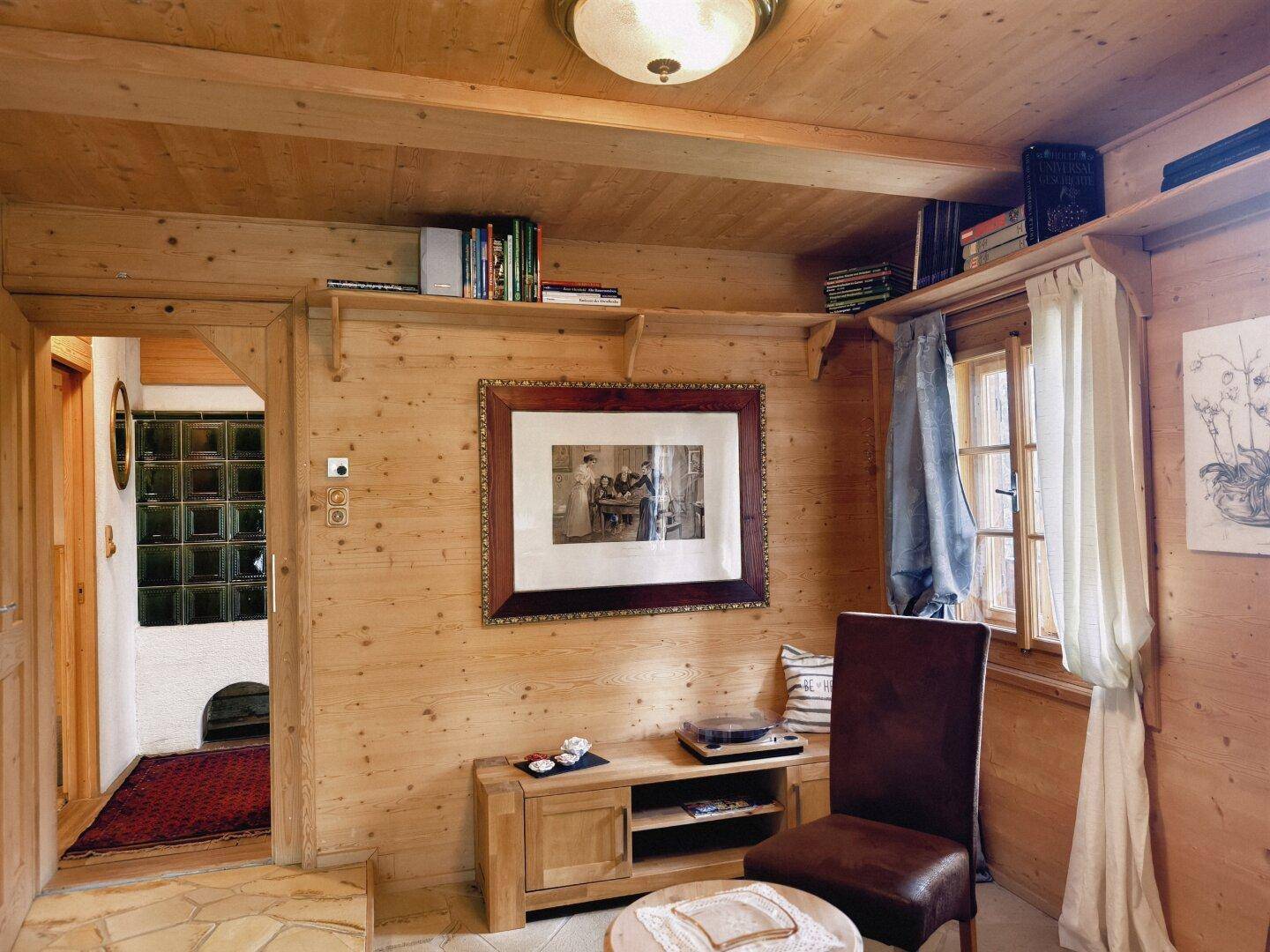 Wohnbereich mit Holzwänden, Bücherregalen und einem traditionellen Kachelofen im Hintergrund.