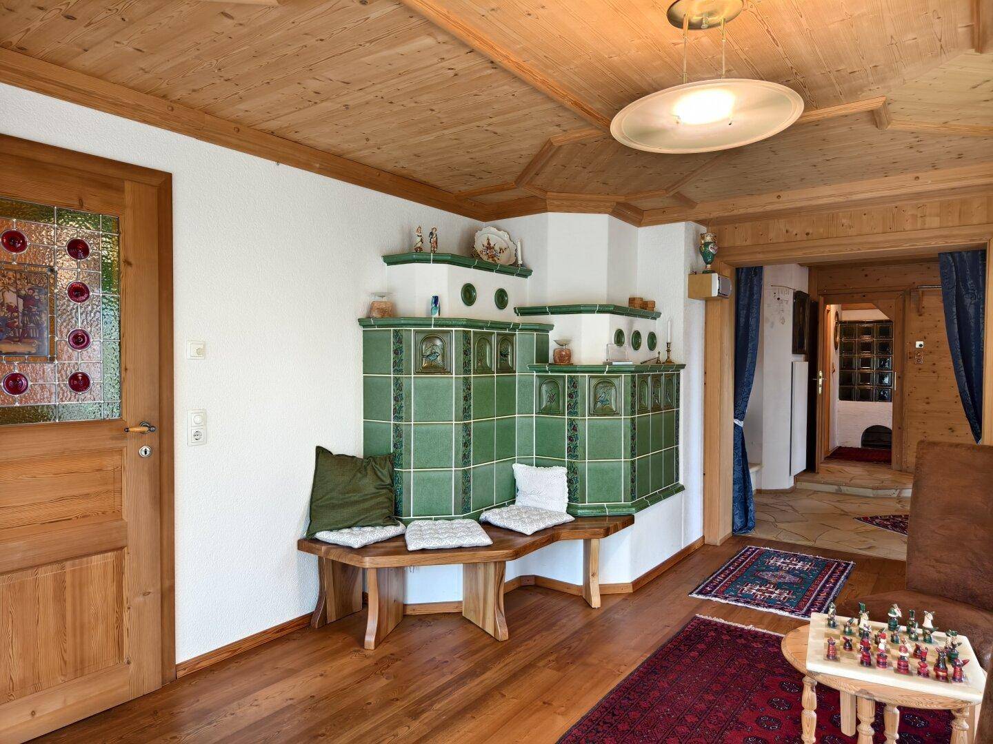 Heller Wohnbereich mit großem Kachelofen, Holzboden und Zugang zu weiteren Räumen.