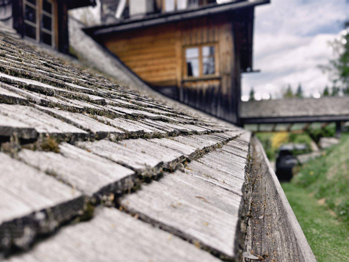 Nahaufnahme eines traditionellen Holzschindeldachs mit Moosbewuchs und rustikaler Holzfassade im Hintergrund.
