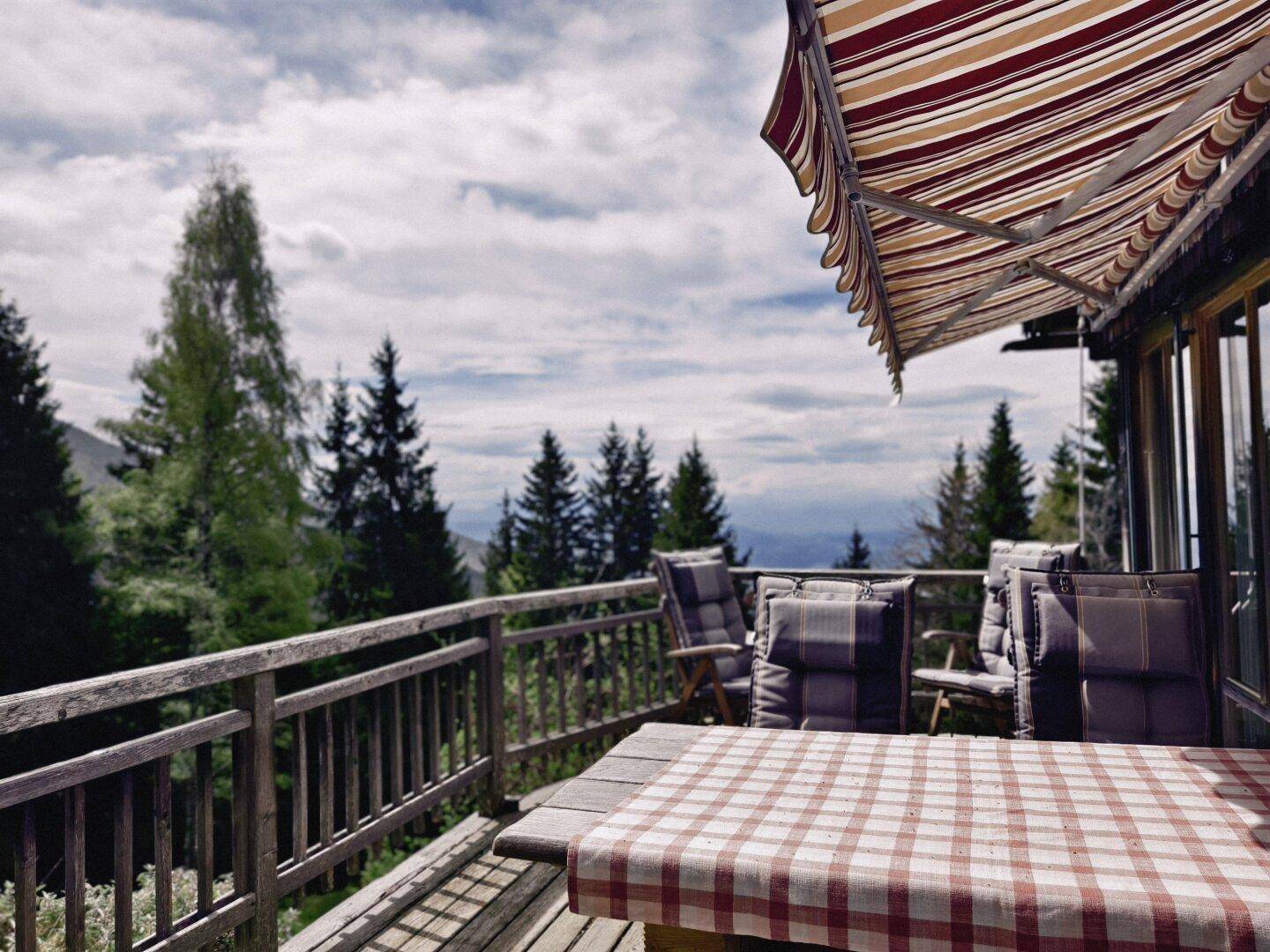 Gemütliche Terrasse mit karierter Tischdecke und Sitzgelegenheiten, umgeben von Nadelbäumen mit Bergblick.