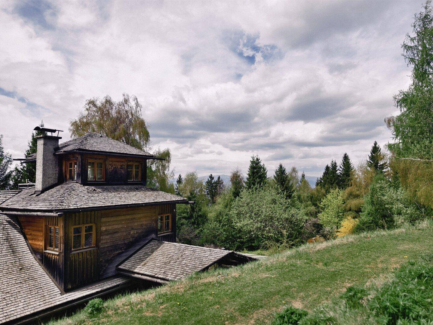 Traditionelles Holzhaus mit mehreren Dächern und einem Schornstein, eingebettet in eine grüne Hügellandschaft.