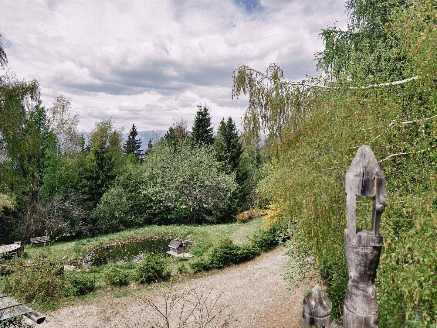 Weitläufiger Garten mit einem natürlichen Teich, umgeben von Bäumen und einer rustikalen Holzskulptur.