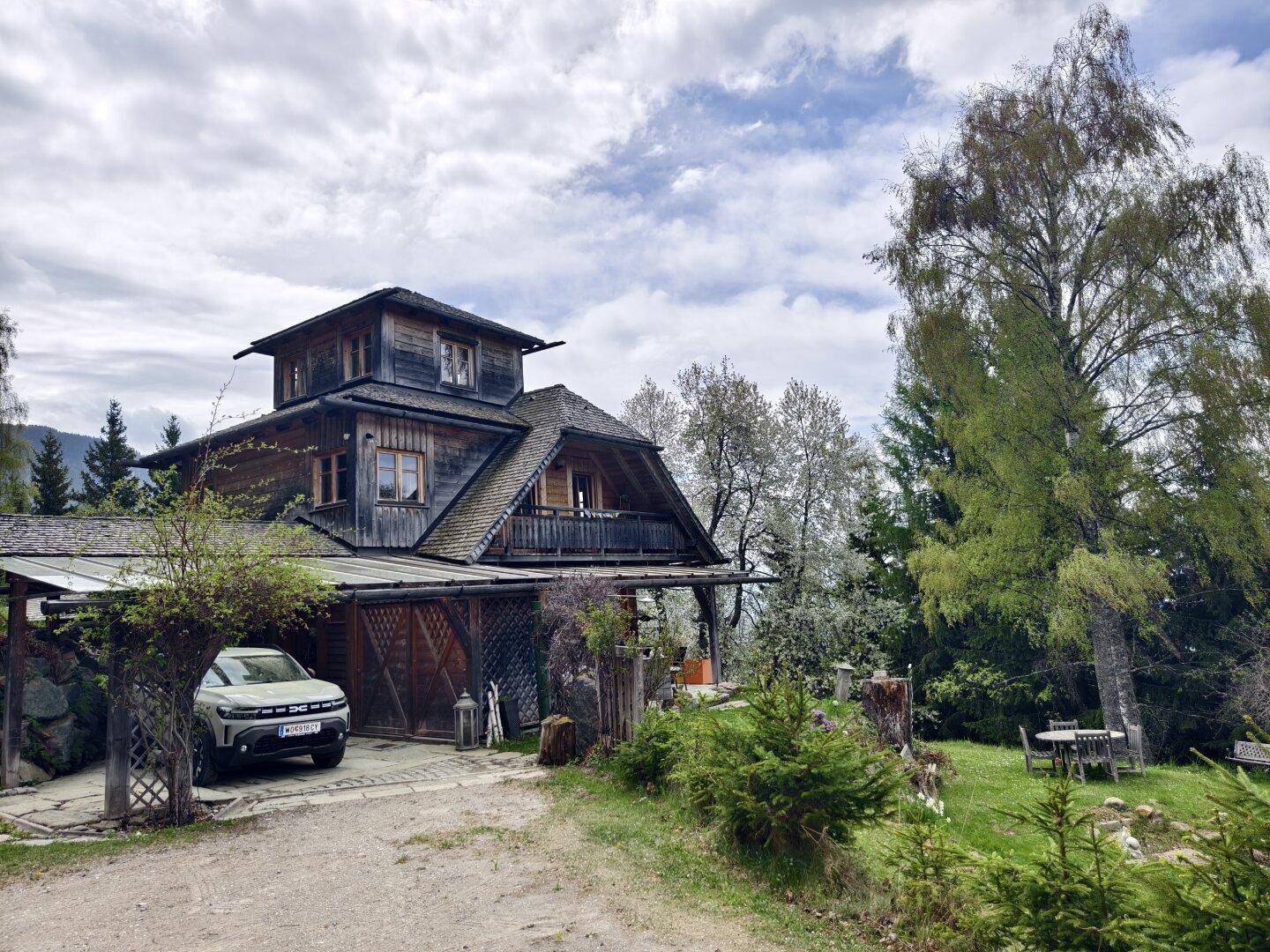 Großes Holzhaus im Chalet-Stil mit Carport und Gartenbereich, umgeben von Bäumen und Natur.