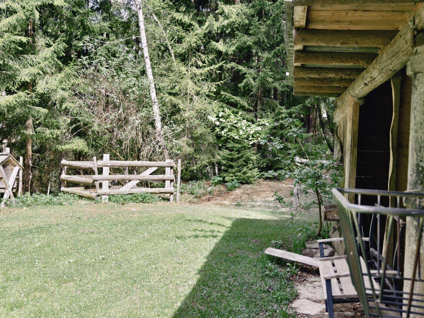 Einfacher Holzschuppen am Rande einer Wiese, mit einem rustikalen Holzzaun und Wald im Hintergrund.