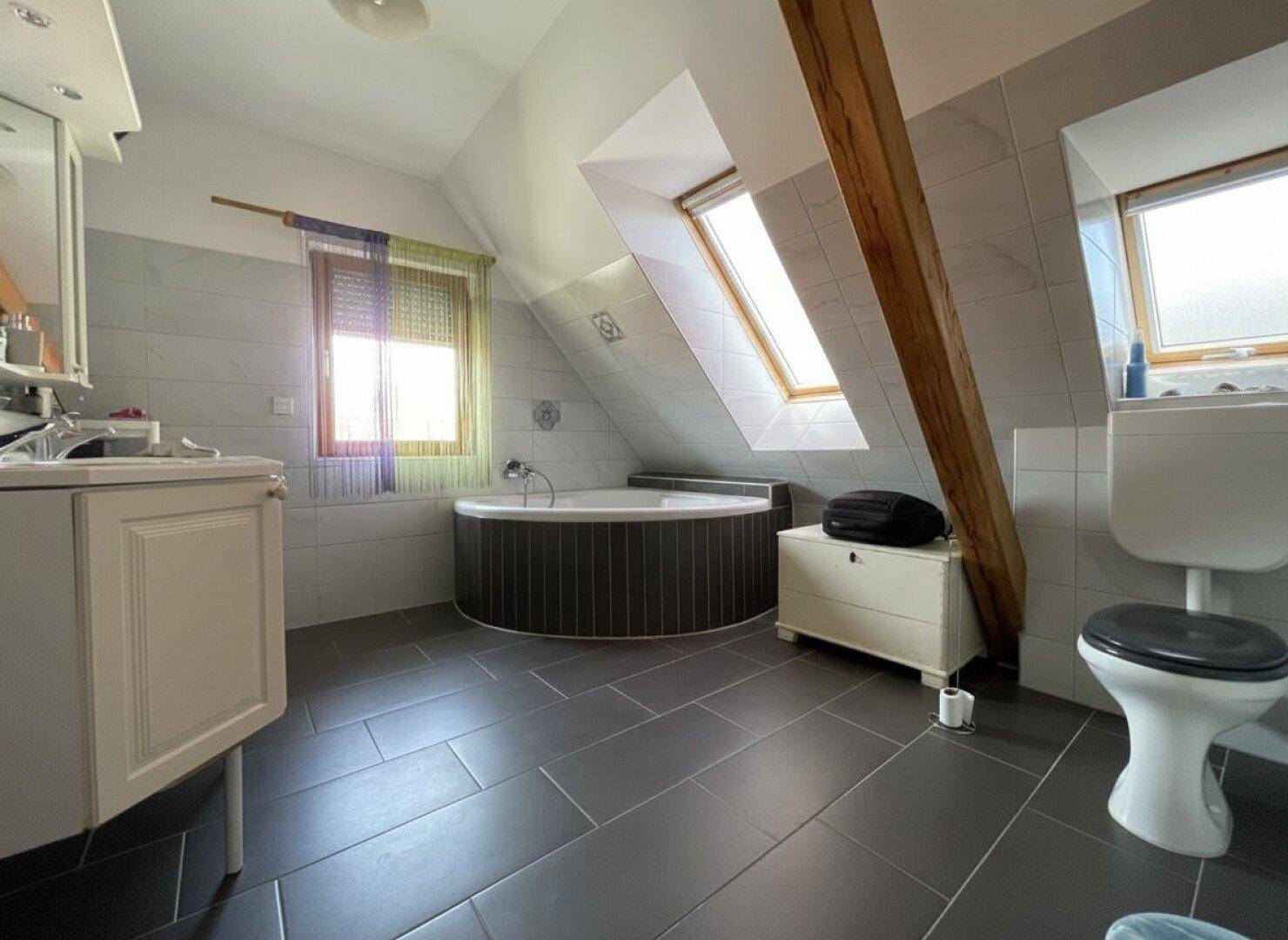 Helles Dachgeschoss-Badezimmer mit Eckbadewanne, Toilette und zwei Fenstern.
