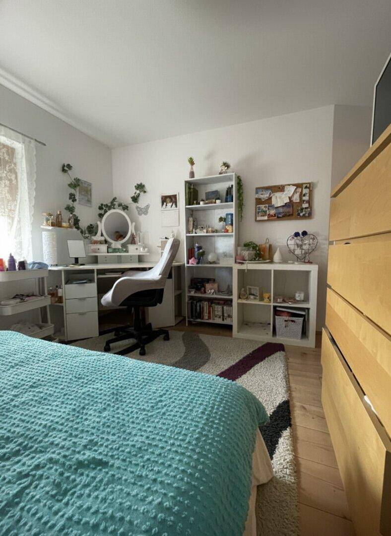 Helles Schlafzimmer im Dachgeschoss mit Schreibtisch, Regal und Teppich auf Laminatboden.