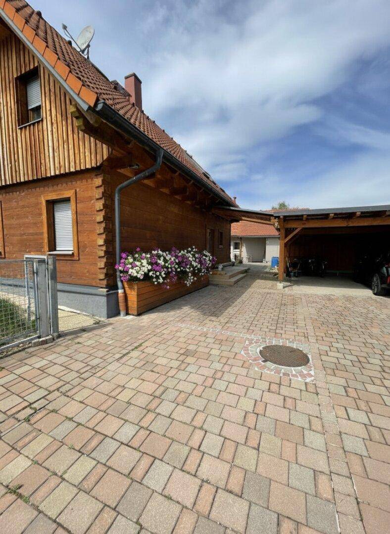 Seitenansicht des Holzhauses mit gepflasterter Einfahrt und Blumenkästen unter den Fenstern.