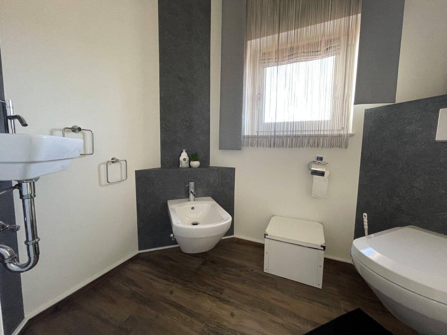 Modernes WC mit Bidet, wandhängender Toilette und Fenster, akzentuiert durch dunkle Wandpaneele.