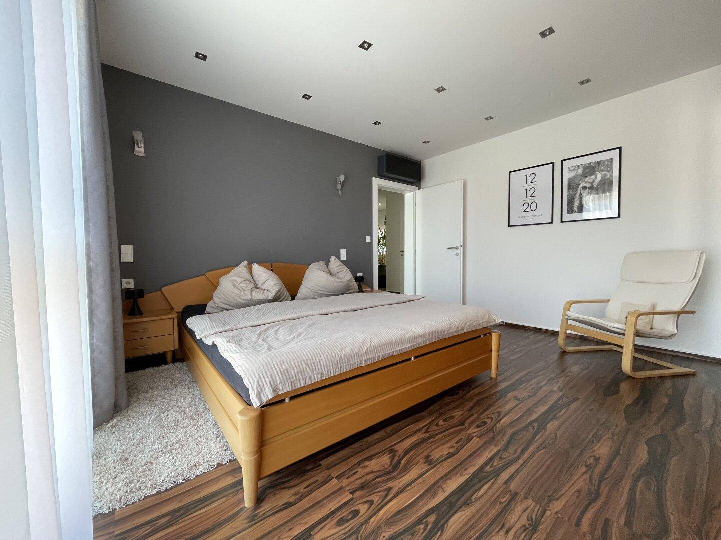 Helles Schlafzimmer mit Holzbett, einer grauen Akzentwand und einem bequemen weißen Sessel.