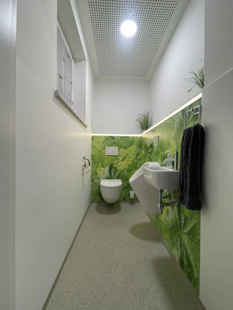 Modernes WC mit Urinal, Toilette und kleinem Waschbecken, akzentuiert durch eine grüne Wandgestaltung.