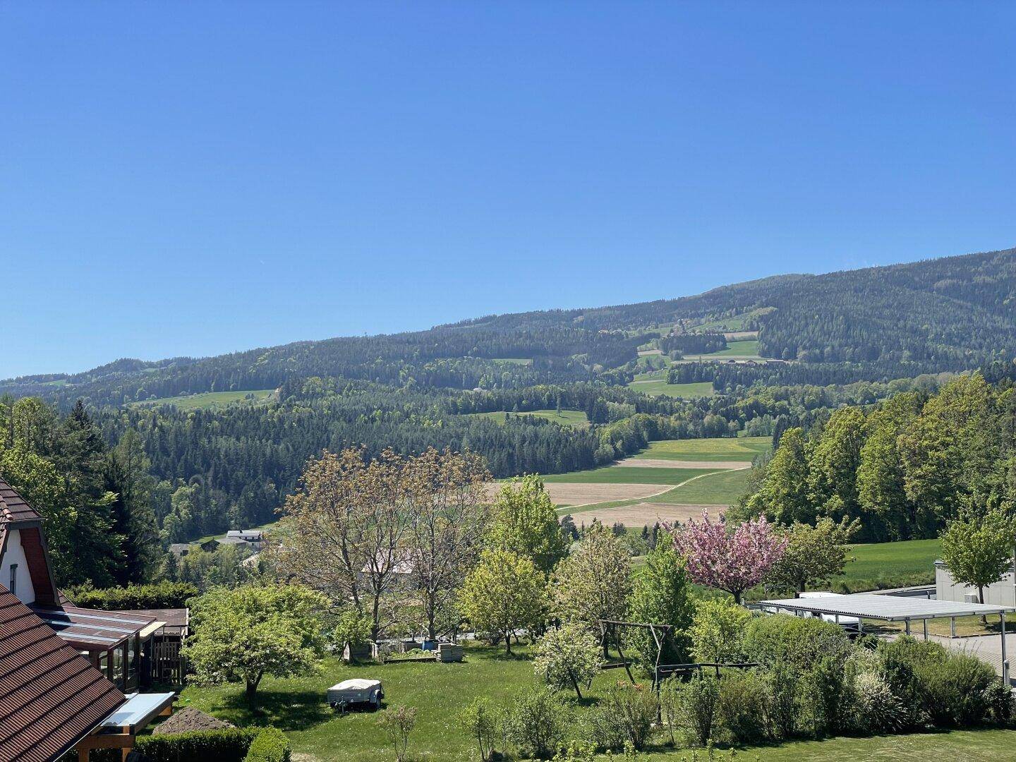 Malerischer Ausblick auf eine grüne Landschaft mit Häusern, Bäumen und sanften Hügeln im Hintergrund.