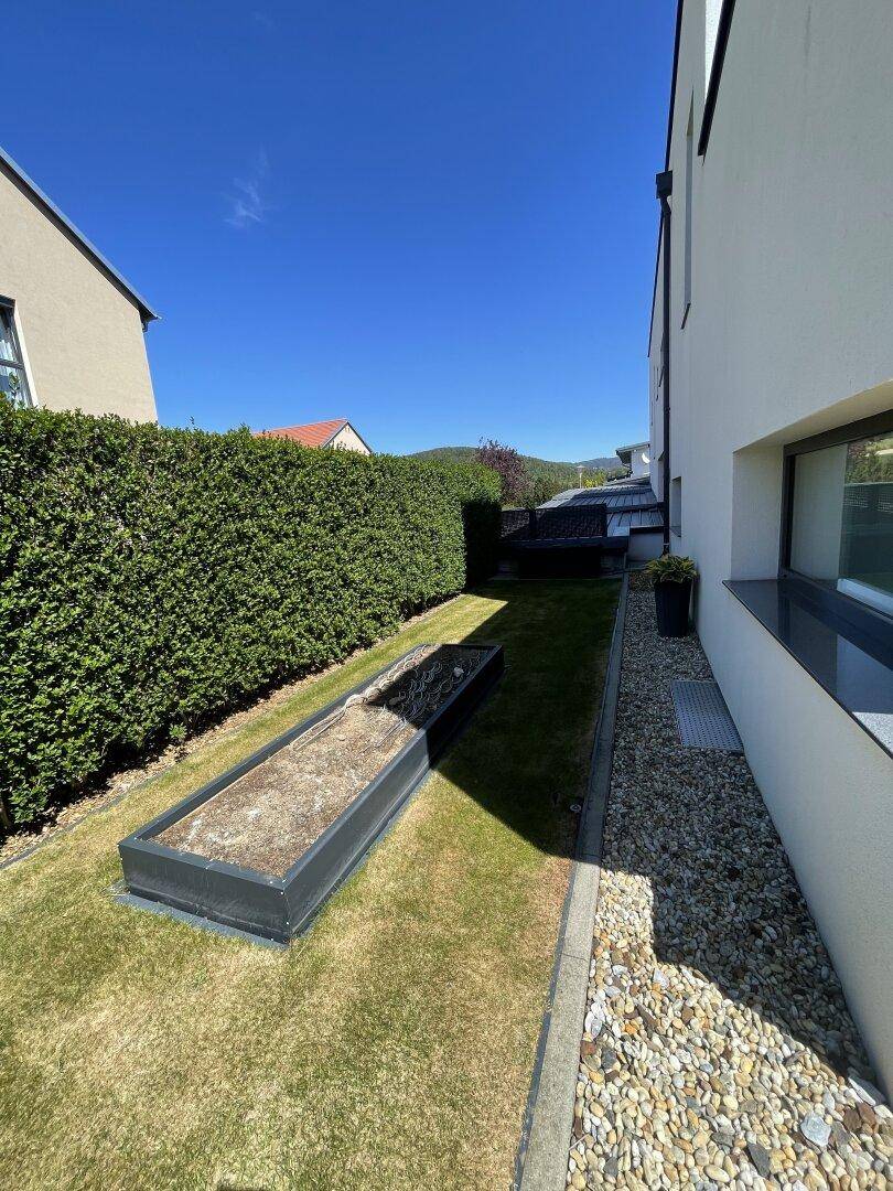 Seitlicher Gartenbereich mit Rasenfläche, einem erhöhten Pflanzbeet, Kiesweg und einer gepflegten Hecke.