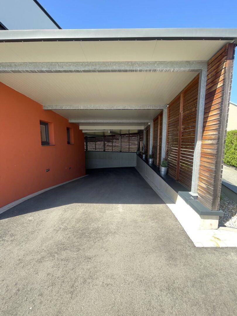 Überdachter Carport mit Holzlamellen und direkter Zufahrt, angrenzend an die orangefarbene Hausfassade.
