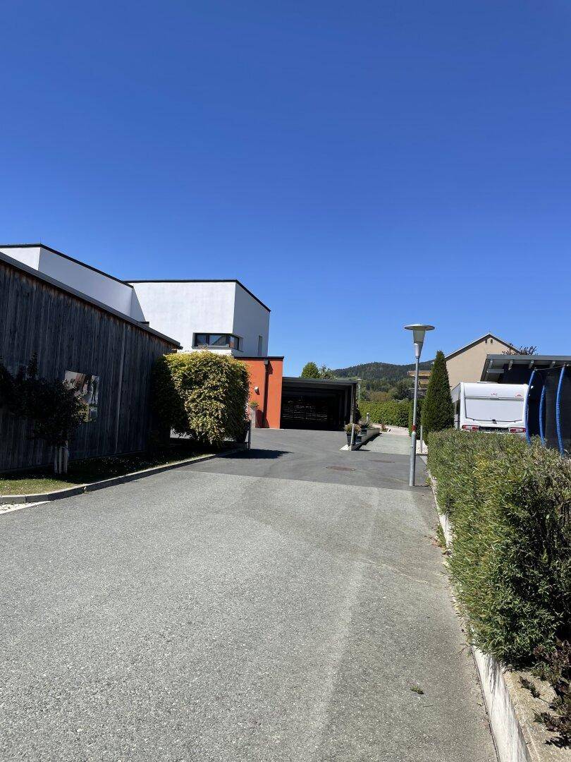 Zufahrt zu einem modernen Haus mit Carport, umgeben von gepflegten Hecken und weiteren Gebäuden.