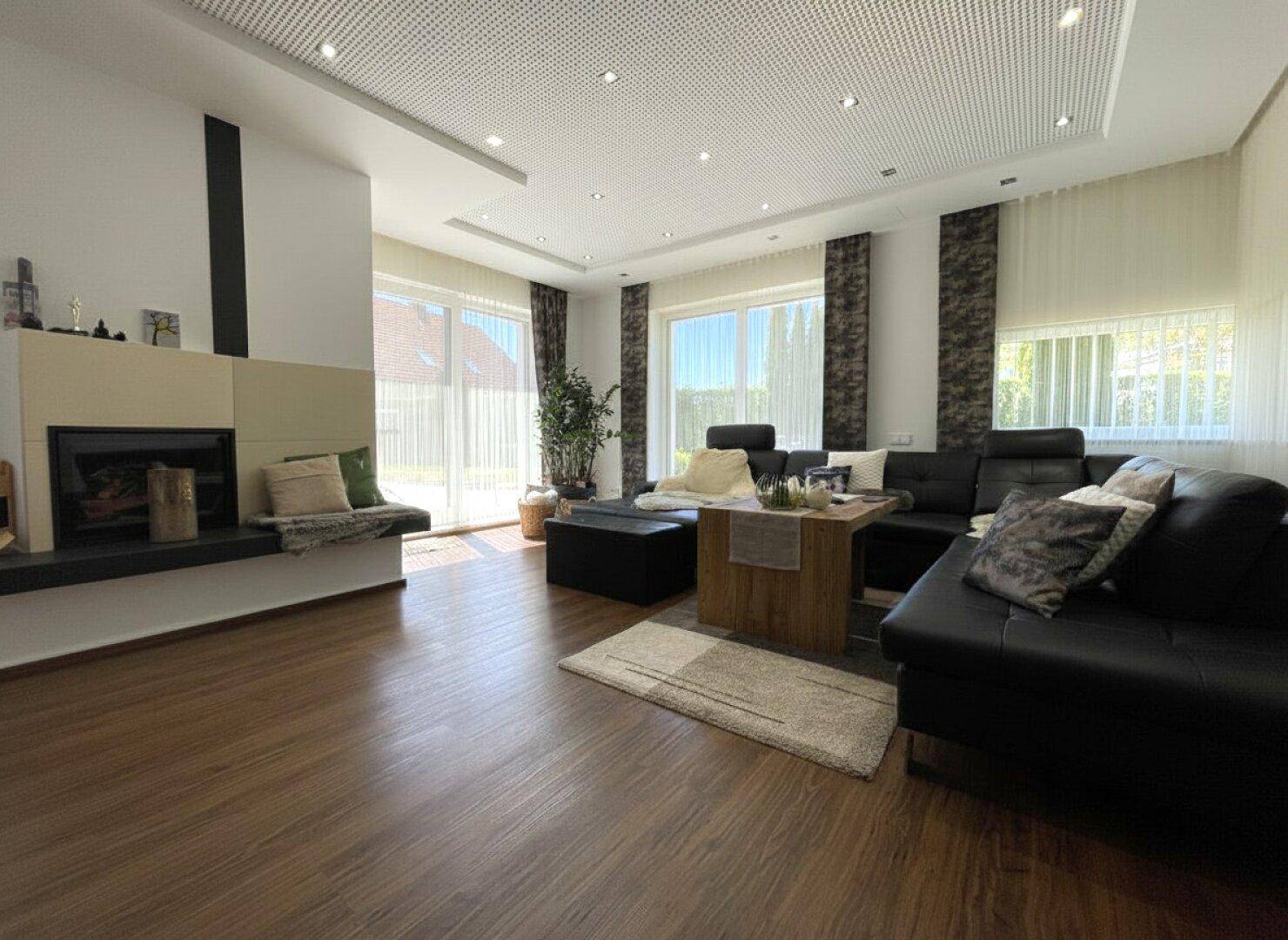 Helles Wohnzimmer mit großen Fenstern, Holzboden und gemütlicher Sitzecke am Kamin.