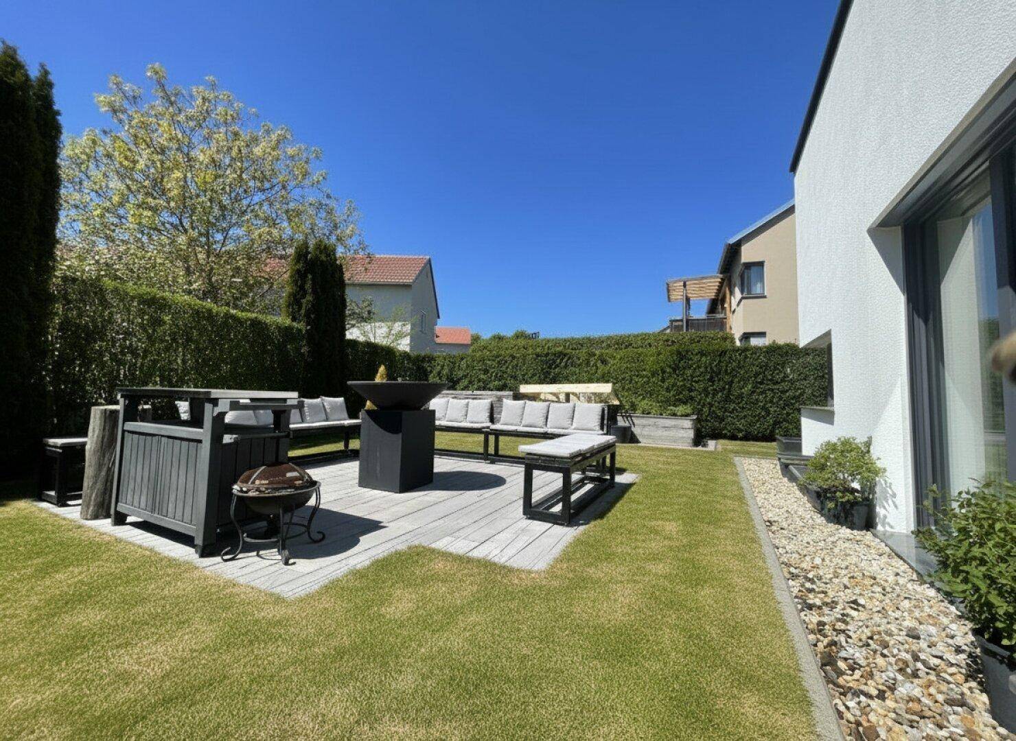 Gemütlicher Außenbereich mit Holzterrasse, modernen Gartenmöbeln, Rasenfläche und hoher Hecke.