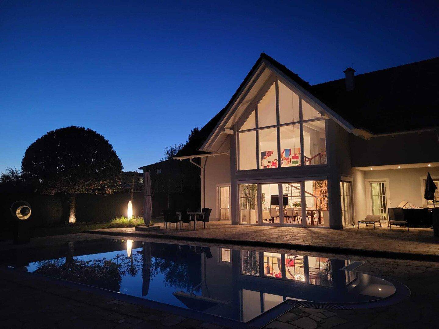 Beleuchteter Garten mit Pool und reflektierendem Wasser bei Nacht, das Haus ist ebenfalls beleuchtet.