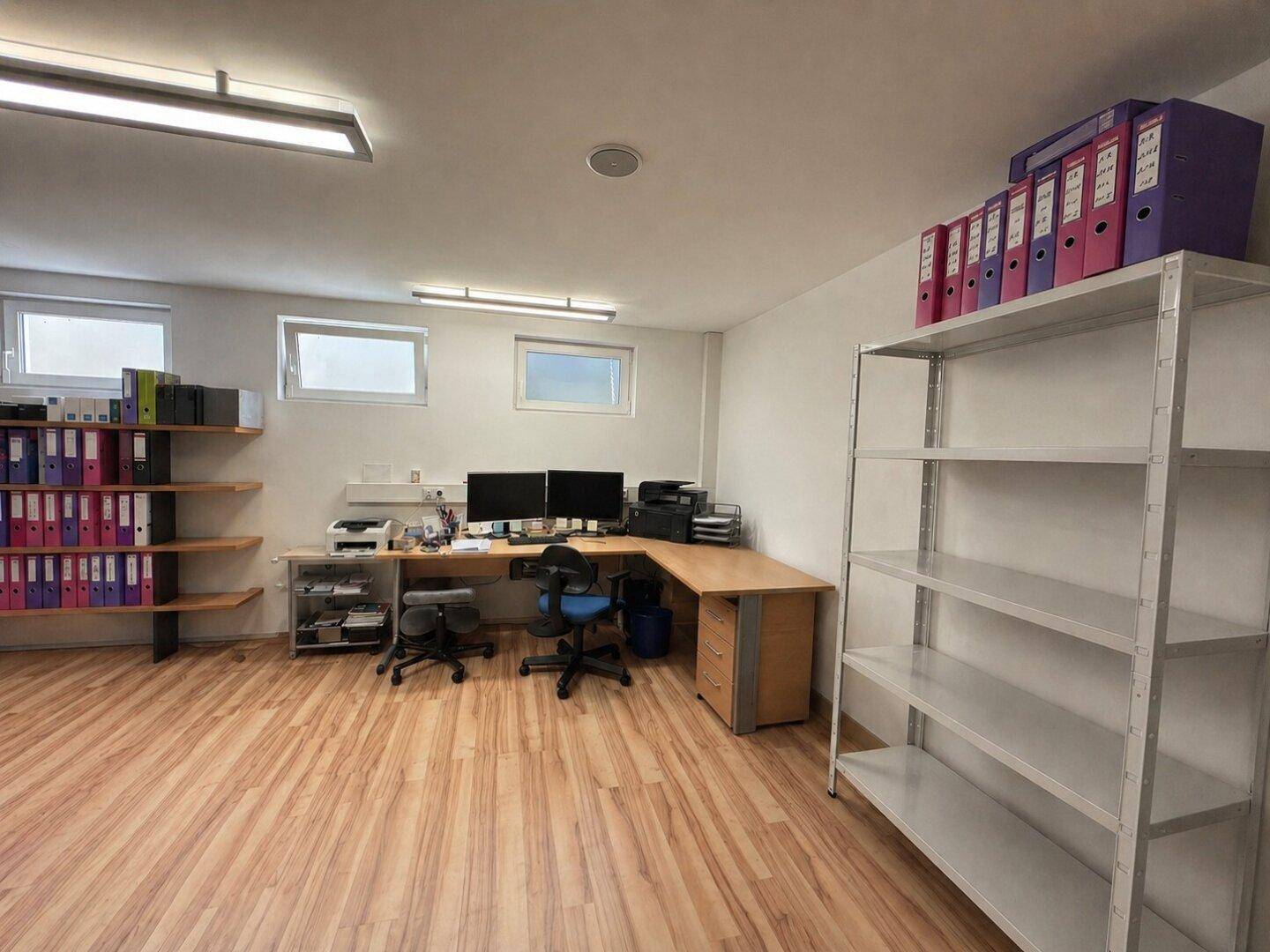 Geräumiges Büro im Untergeschoss mit mehreren Arbeitsplätzen, Regalen und Laminatboden.