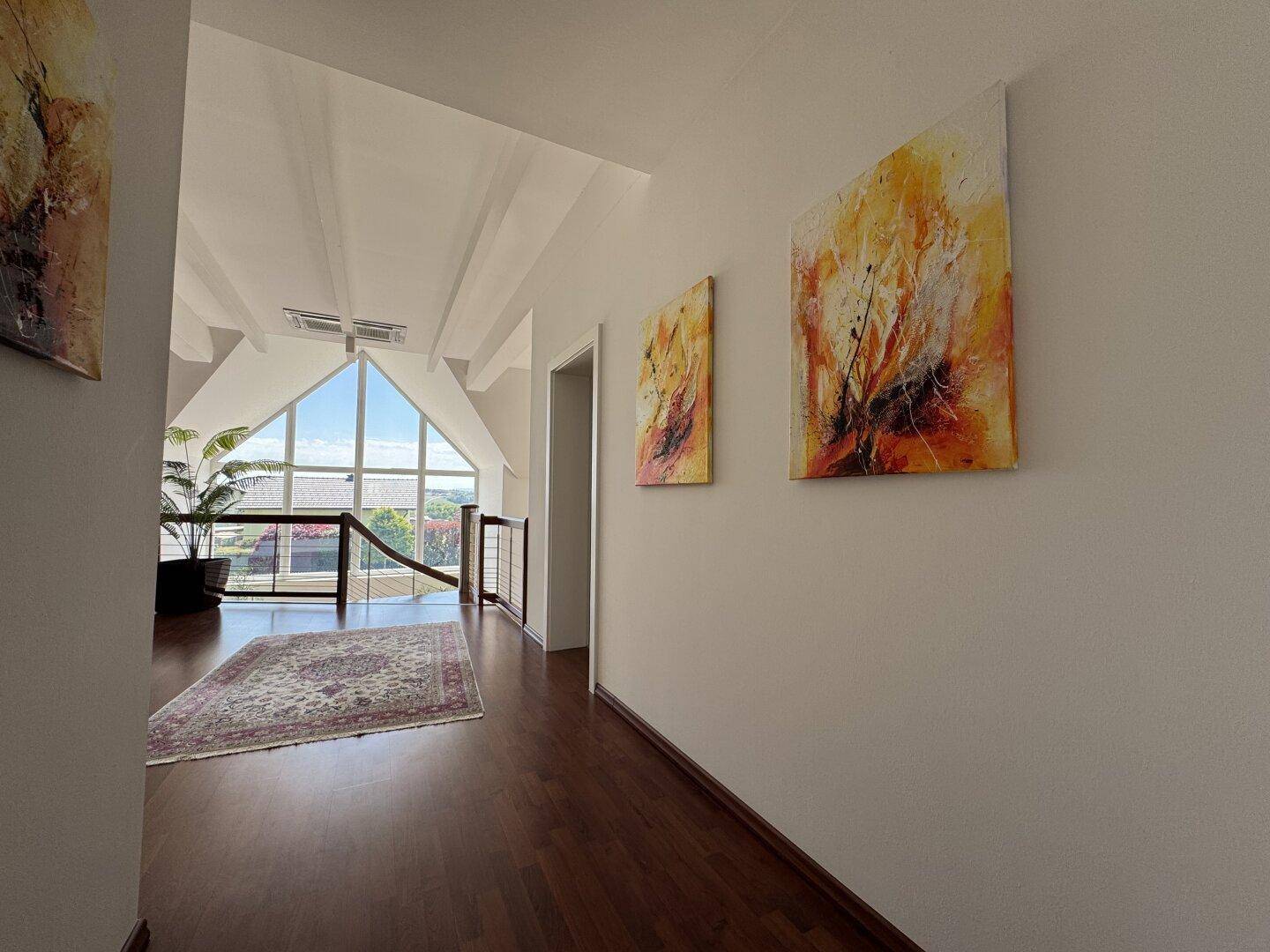 Geräumiger Flur im Dachgeschoss mit Holzboden, Kunstwerken und Blick durch die Giebelfenster.