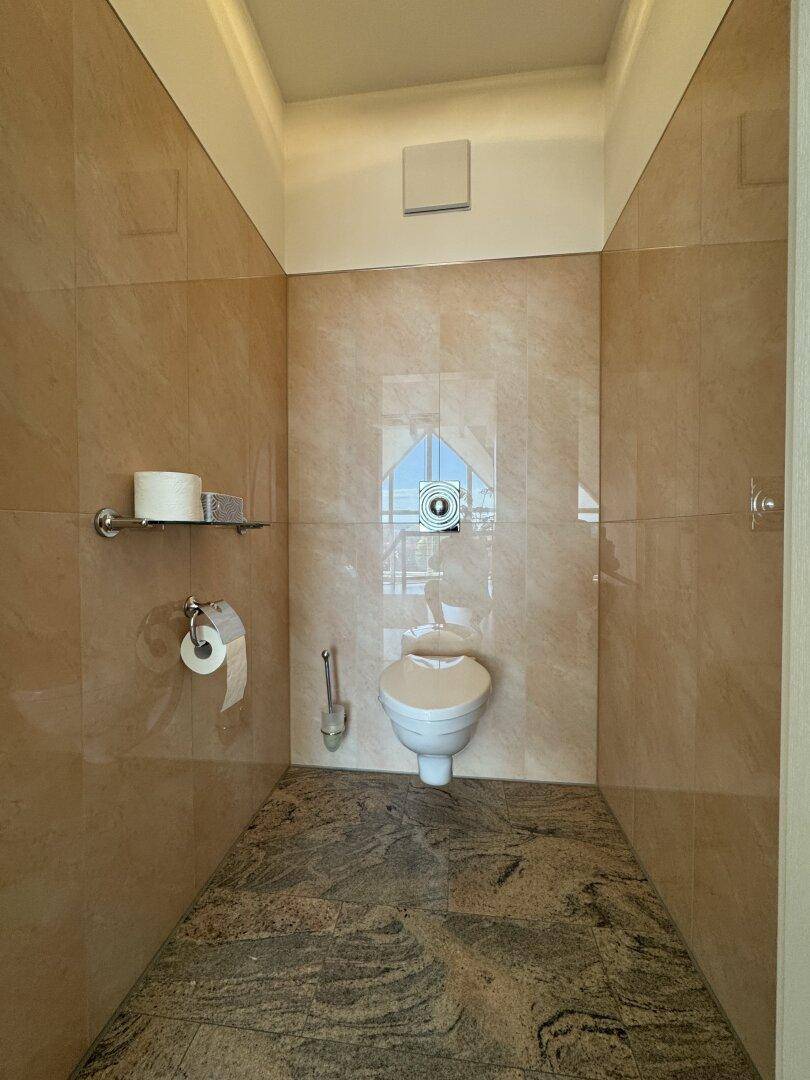Separate Toilette mit Wand-WC und beigen Fliesen, ausgestattet mit einem Fenster.