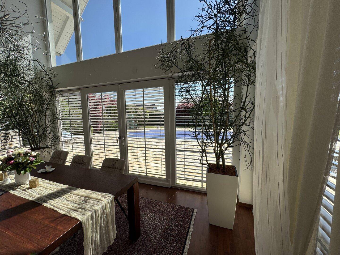 Essbereich mit Blick auf den Pool und den Garten durch große Fenster mit Jalousien.