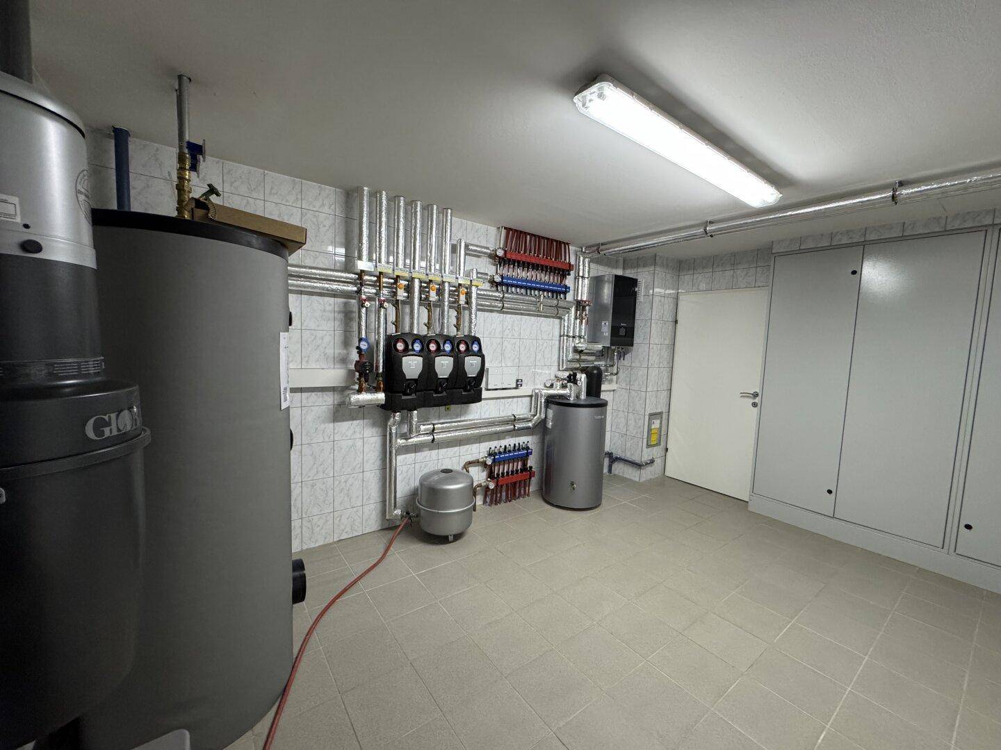 Heizungsraum mit großen Warmwasserspeichern, Verteilern und Rohrleitungen, gefliesten Wänden.