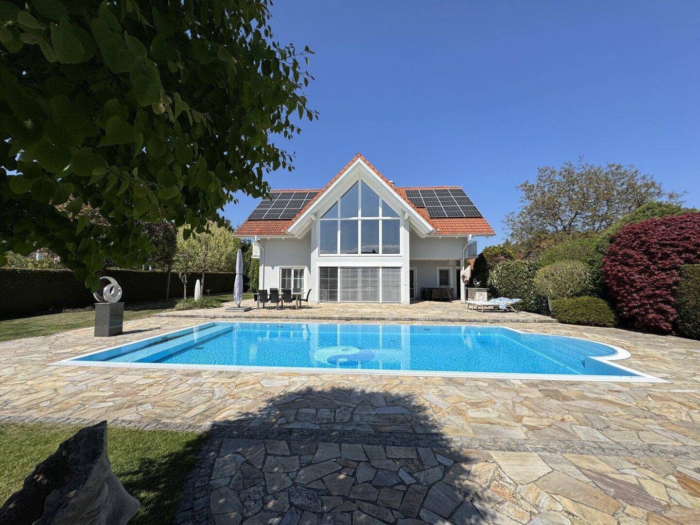 Frontansicht des Hauses mit großem Pool und weitläufiger Steinterrasse unter blauem Himmel.