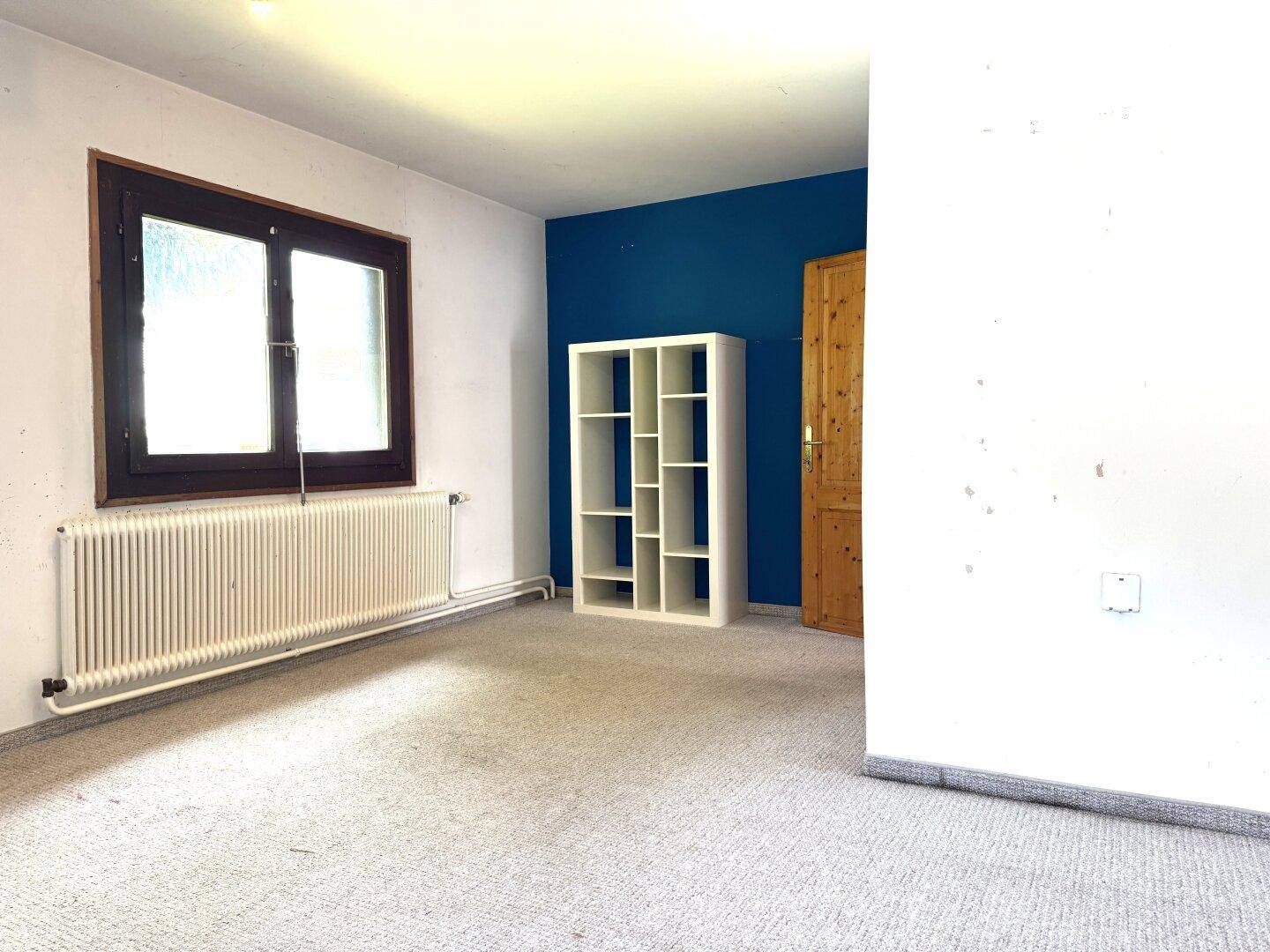 Zimmer mit Teppichboden, blauer Akzentwand und großem Fenster, ideal als Kinder- oder Gästezimmer.