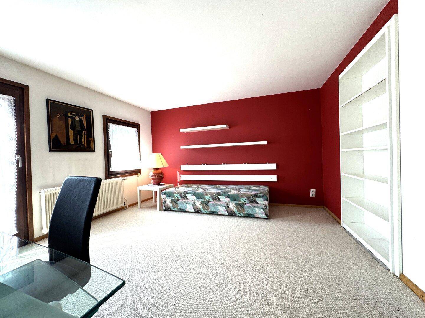 Vielseitiger Raum mit roter Akzentwand, Teppichboden und großem Fenster, ideal als Büro oder Gästezimmer.