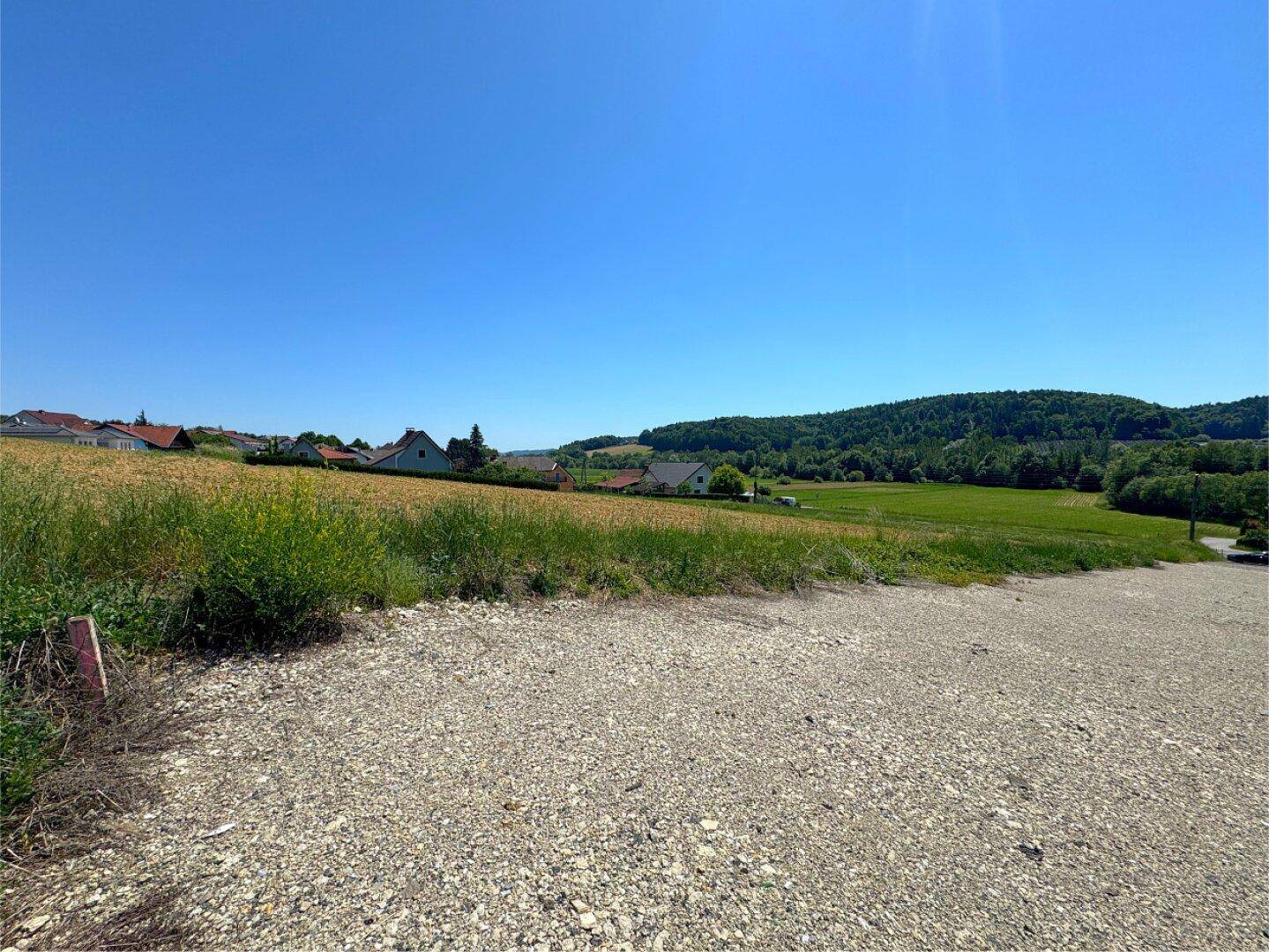 Schotterfläche im Vordergrund, übergehend in grüne Wiesen mit ländlicher Bebauung am Horizont.