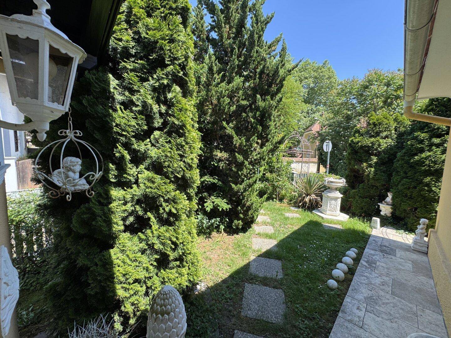 Blick in den grünen Garten mit hohen Bäumen und dekorativen Gartenfiguren.
