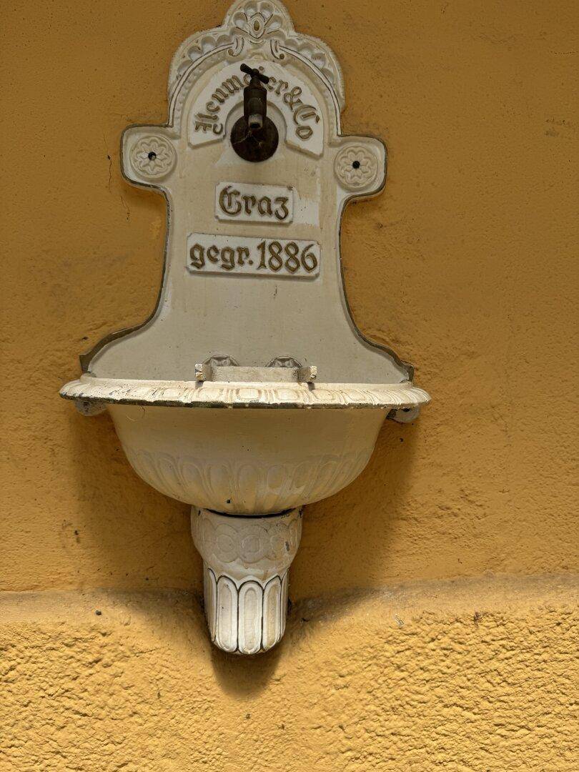 Historischer Wasserhahn mit Inschrift "Graz gegr. 1886" an der gelben Hauswand.