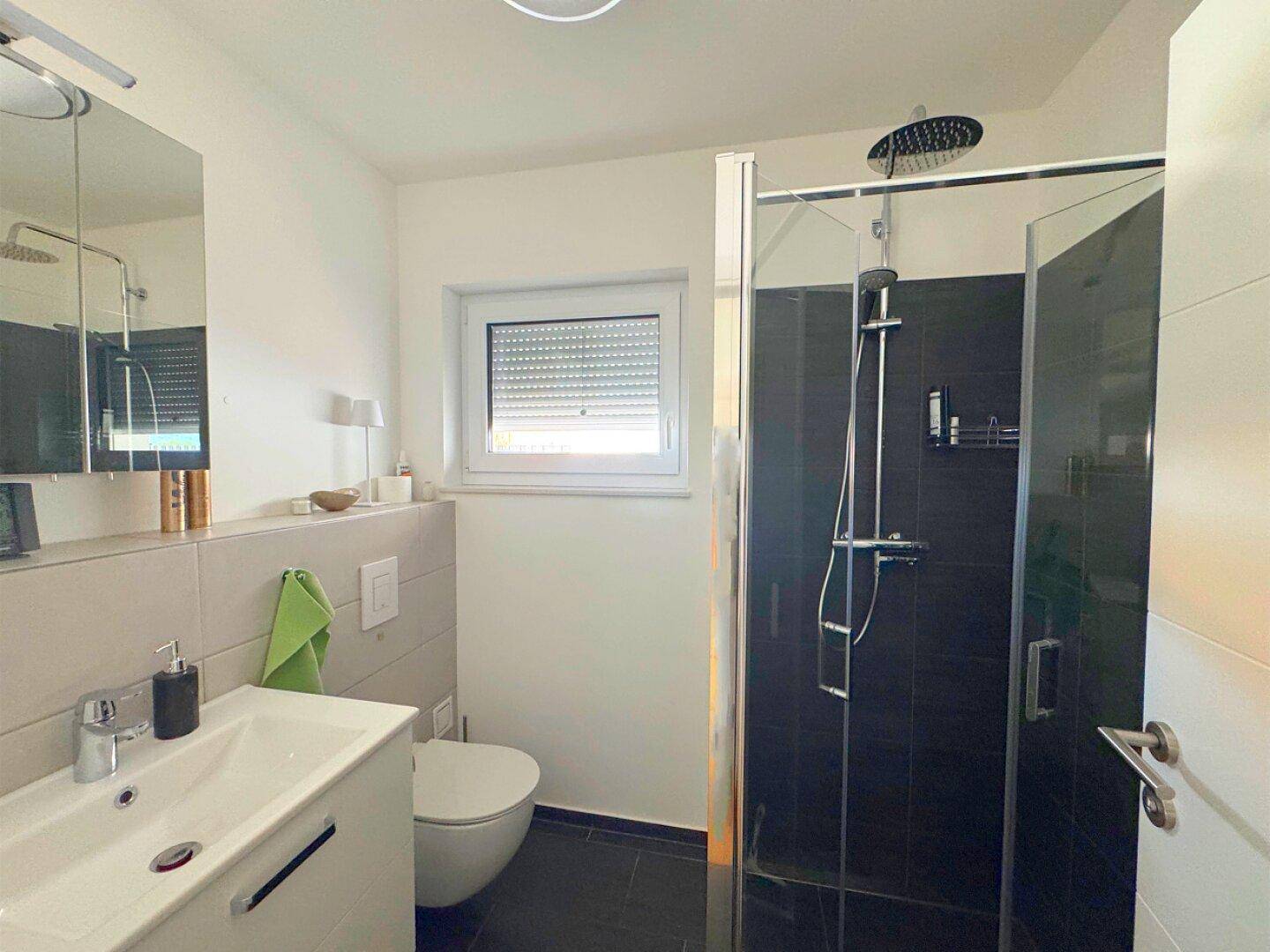 Helles Badezimmer mit Dusche, Waschbecken und Fenster mit Rollladen.