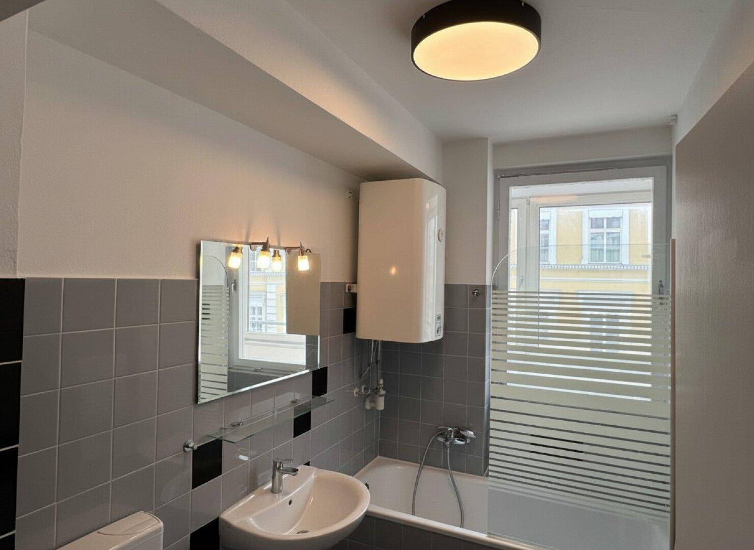 Badezimmer mit Badewanne, Waschbecken, Spiegel und Fenster, ausgestattet mit Boiler und grauen Fliesen.