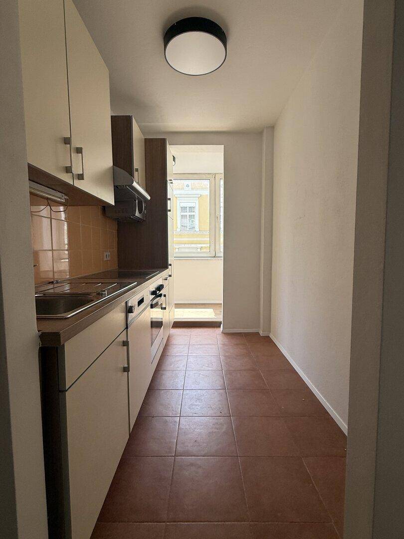 Schmale Küche mit Einbauschränken, Herd und Blick durch das Fenster auf die Straße, funktional eingerichtet.