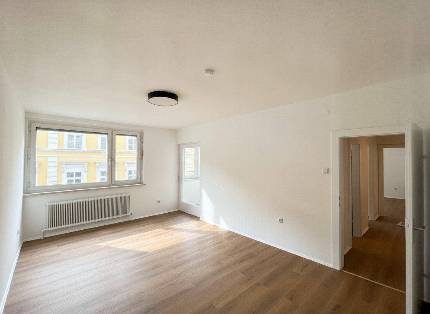 Leeres Zimmer mit Holzboden, großen Fenstern und Zugang zu einem möglichen Balkon, bietet viel Tageslicht.
